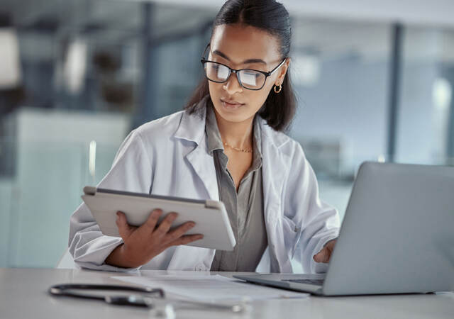 Doctor reading a tablet while working on a laptop