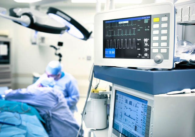 A closeup of operating room monitors while a surgeon works on a patient in the background