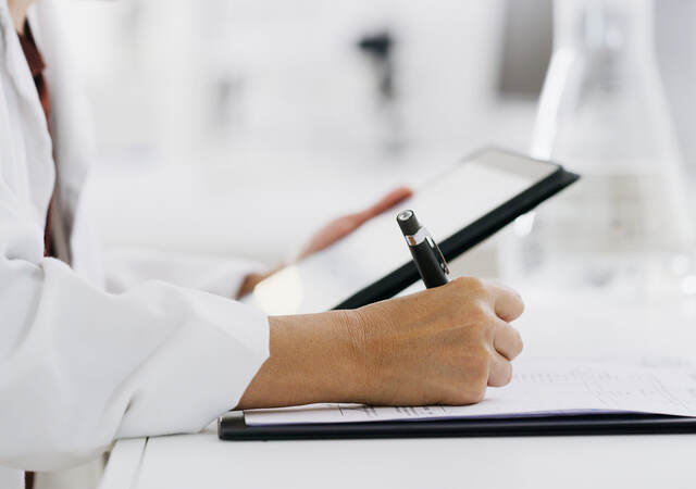 Closeup of a medical professional reading a tablet and making notes on paper while standing in a lab