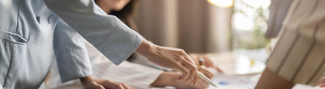 A closeup of a person using a stylus on a tablet on a desk with other people collaborating around them