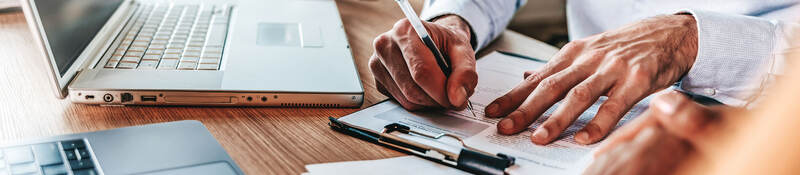 Person sitting at desk with a laptop taking notes on a clipboard