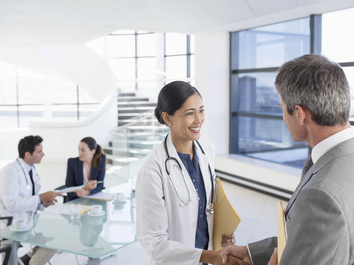 A doctor shaking hands with a business professional while another doctor has a meeting in the background