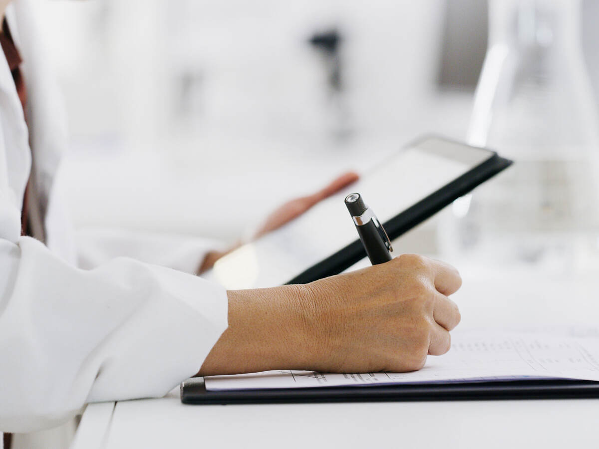 Closeup of a medical professional reading a tablet and making notes on paper while standing in a lab